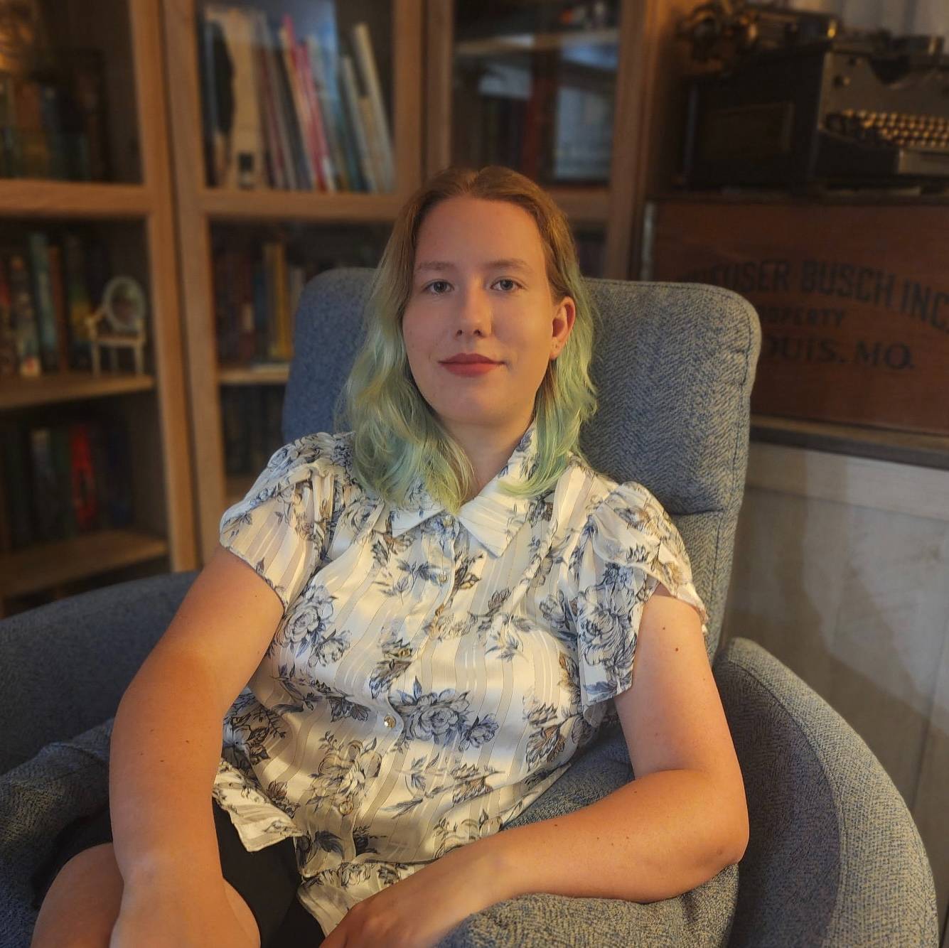 Anneli is wearing a black and grey floral button up, has green ombre hair, and is wearing a bright read lipstick. 