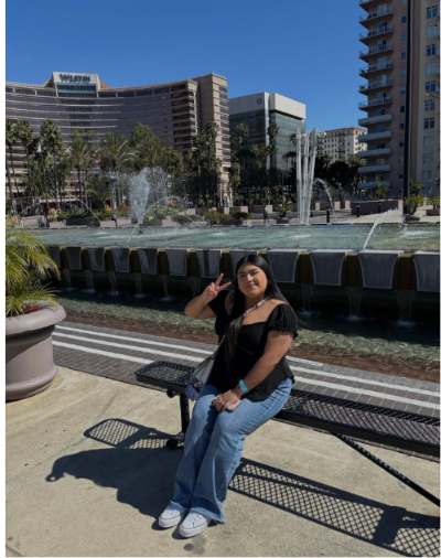 Ximena is sitting next to a fountain. She is wearing a black shirt and jeans. She is throwing up a peace sign. 