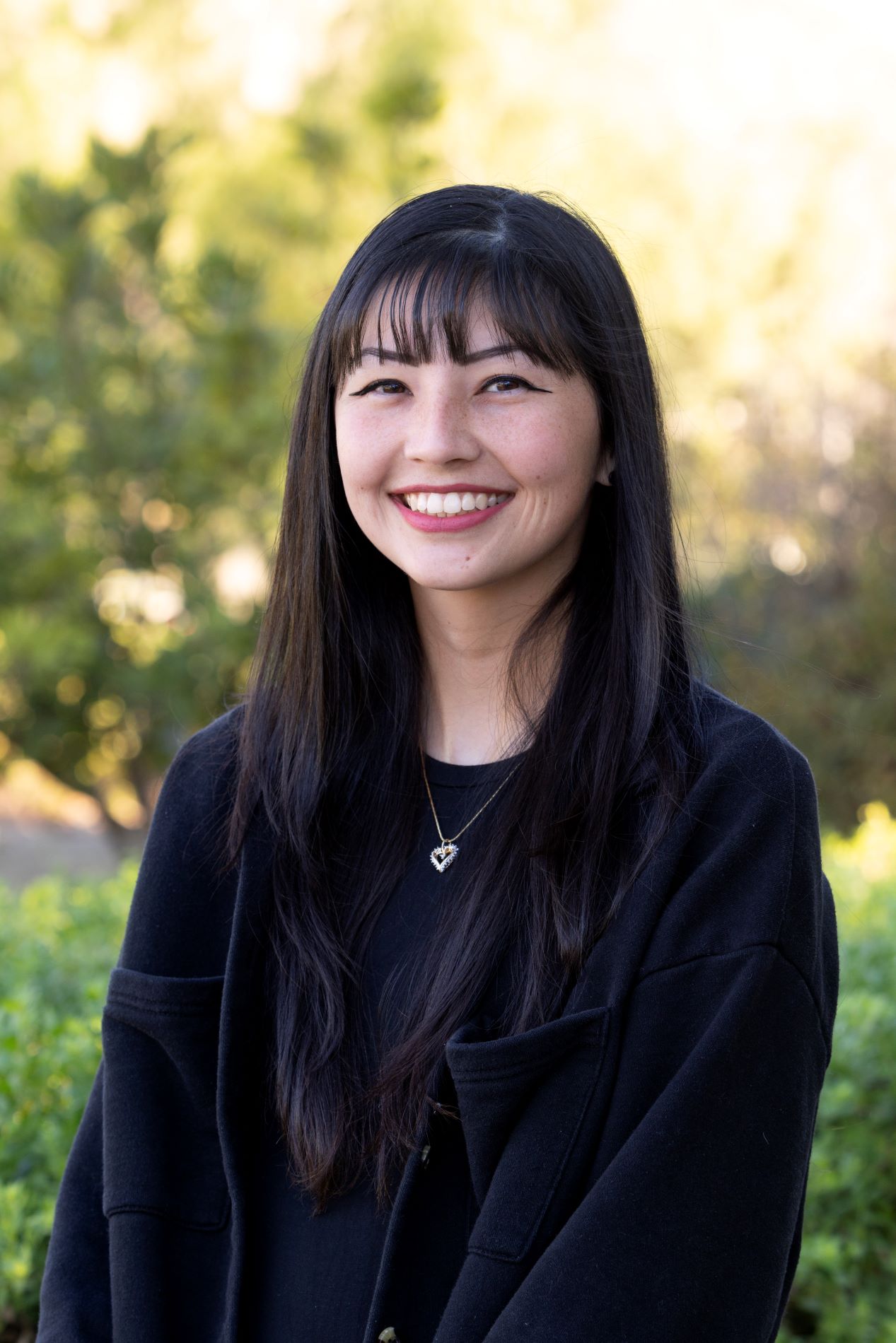 A woman with dark hair wearing a black jacket is smiling at the camera in front of a green foliage background. 